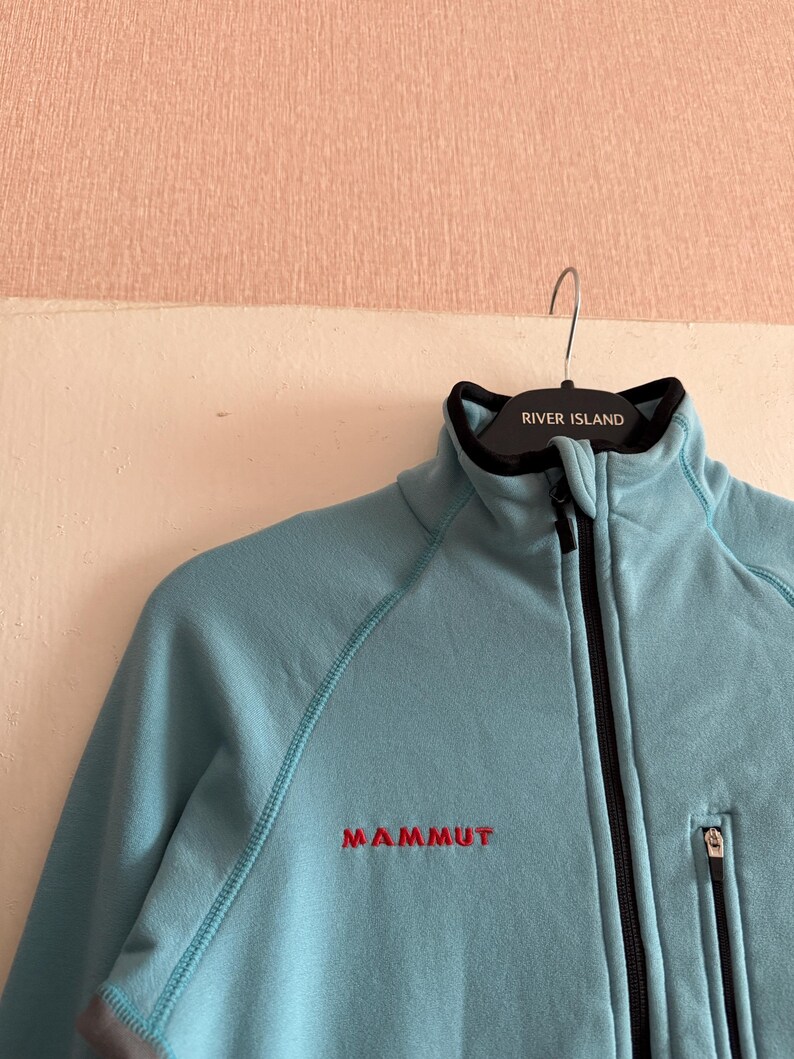 Puede incluir: Chaqueta polar azul claro con cremallera negra y un peque&ntilde;o bolsillo con cremallera. La chaqueta tiene la palabra "MAMMUT" bordada en rojo en el pecho. El cuello es negro y la chaqueta est&aacute; colgada en una percha con "RIVER ISLAND".