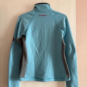 Puede incluir: Chaqueta azul claro con cremallera, cuello alto y mangas largas. La chaqueta tiene paneles laterales grises y la palabra "MAMMUT" en letras rojas en la parte superior de la espalda. Los pu&ntilde;os y el cuello son negros.