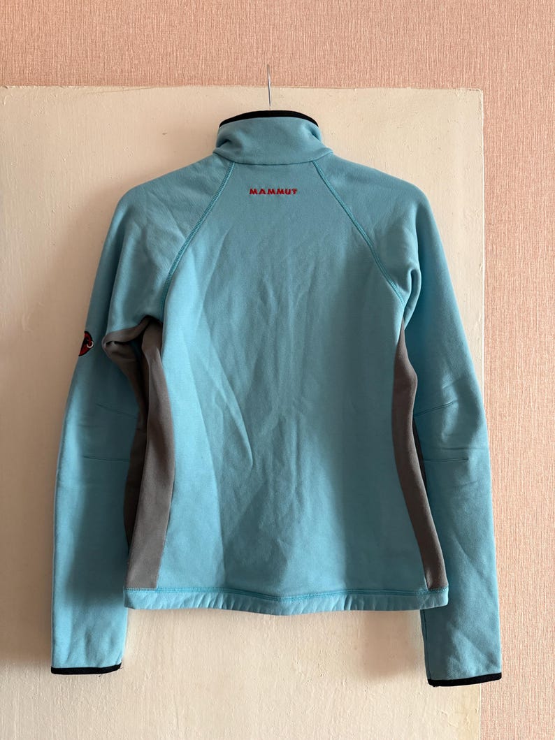 Puede incluir: Chaqueta azul claro con cremallera, cuello alto y mangas largas. La chaqueta tiene paneles laterales grises y la palabra "MAMMUT" en letras rojas en la parte superior de la espalda. Los pu&ntilde;os y el cuello son negros.
