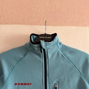 Puede incluir: Chaqueta polar azul claro con cremallera negra y un peque&ntilde;o bolsillo con cremallera. La chaqueta tiene la palabra "MAMMUT" bordada en rojo en el pecho. El cuello es negro con el texto "RIVER ISLAND".