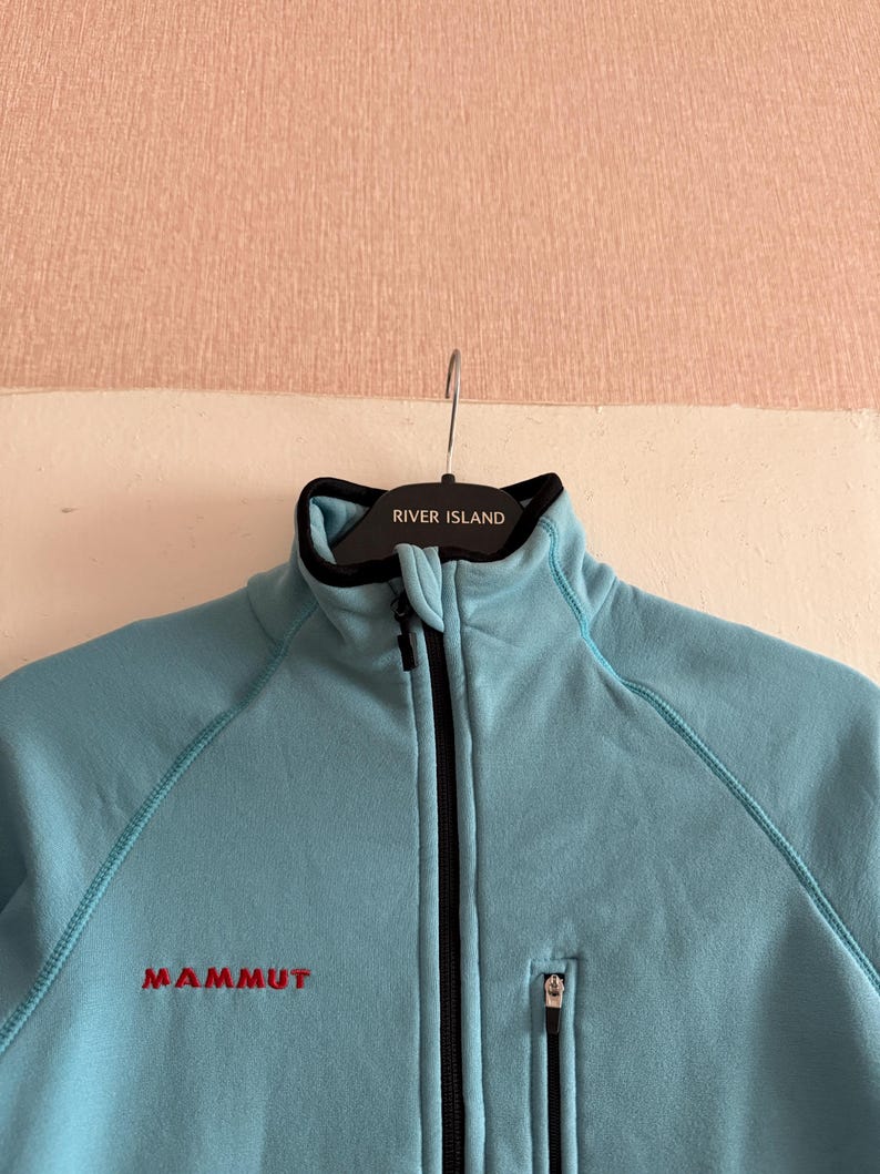 Puede incluir: Chaqueta polar azul claro con cremallera negra y un peque&ntilde;o bolsillo con cremallera. La chaqueta tiene la palabra "MAMMUT" bordada en rojo en el pecho. El cuello es negro con el texto "RIVER ISLAND".