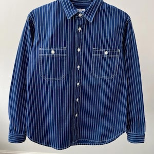Può includere: Una camicia blu a maniche lunghe con sottili gessati bianchi verticali. La camicia ha due tasche sul petto, bottoni bianchi e cuciture bianche. La camicia è appesa a una gruccia nera con la scritta "RIVER ISLAND".