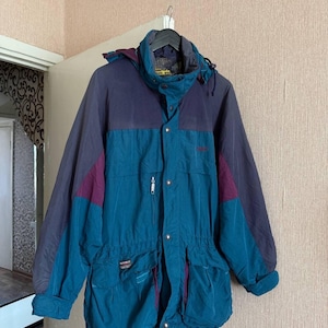 K&ouml;nnte beinhalten: Eine petrolfarbene und dunkelviolette Kapuzenjacke mit hohem Kragen, Frontrei&szlig;verschluss mit Druckkn&ouml;pfen, zwei gro&szlig;en aufgesetzten Taschen mit Klappen, einer kleinen Rei&szlig;verschlusstasche auf der Brust und einem Kordelzug in der Taille.