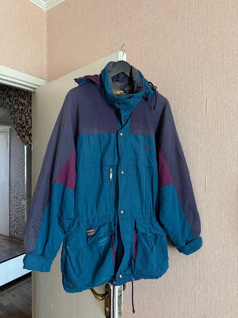 K&ouml;nnte beinhalten: Eine petrolfarbene und dunkelviolette Kapuzenjacke mit hohem Kragen, Frontrei&szlig;verschluss mit Druckkn&ouml;pfen, zwei gro&szlig;en aufgesetzten Taschen mit Klappen, einer kleinen Rei&szlig;verschlusstasche auf der Brust und einem Kordelzug in der Taille.