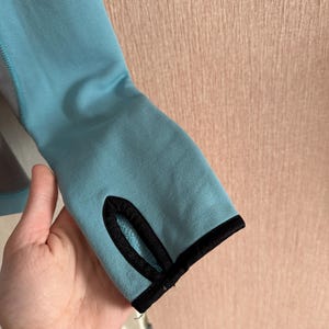 Puede incluir: Primer plano de una manga azul claro con un ribete negro alrededor del orificio para el pulgar. La manga est&aacute; hecha de un material suave y la sostiene una mano. El fondo es una pared rosa claro.