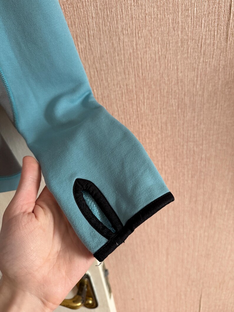 Puede incluir: Primer plano de una manga azul claro con un ribete negro alrededor del orificio para el pulgar. La manga est&aacute; hecha de un material suave y la sostiene una mano. El fondo es una pared rosa claro.