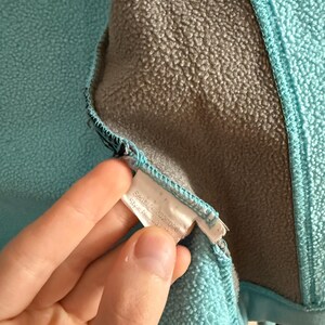 Puede incluir: Primer plano del interior de una chaqueta de forro polar turquesa con forro gris. Se ve una etiqueta blanca con texto. La chaqueta tiene una superficie texturizada y un ribete azul.