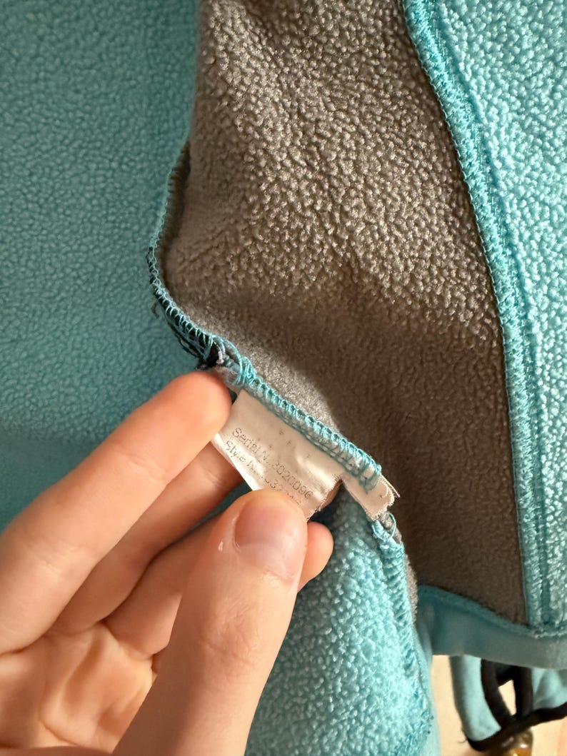 Puede incluir: Primer plano del interior de una chaqueta de forro polar turquesa con forro gris. Se ve una etiqueta blanca con texto. La chaqueta tiene una superficie texturizada y un ribete azul.