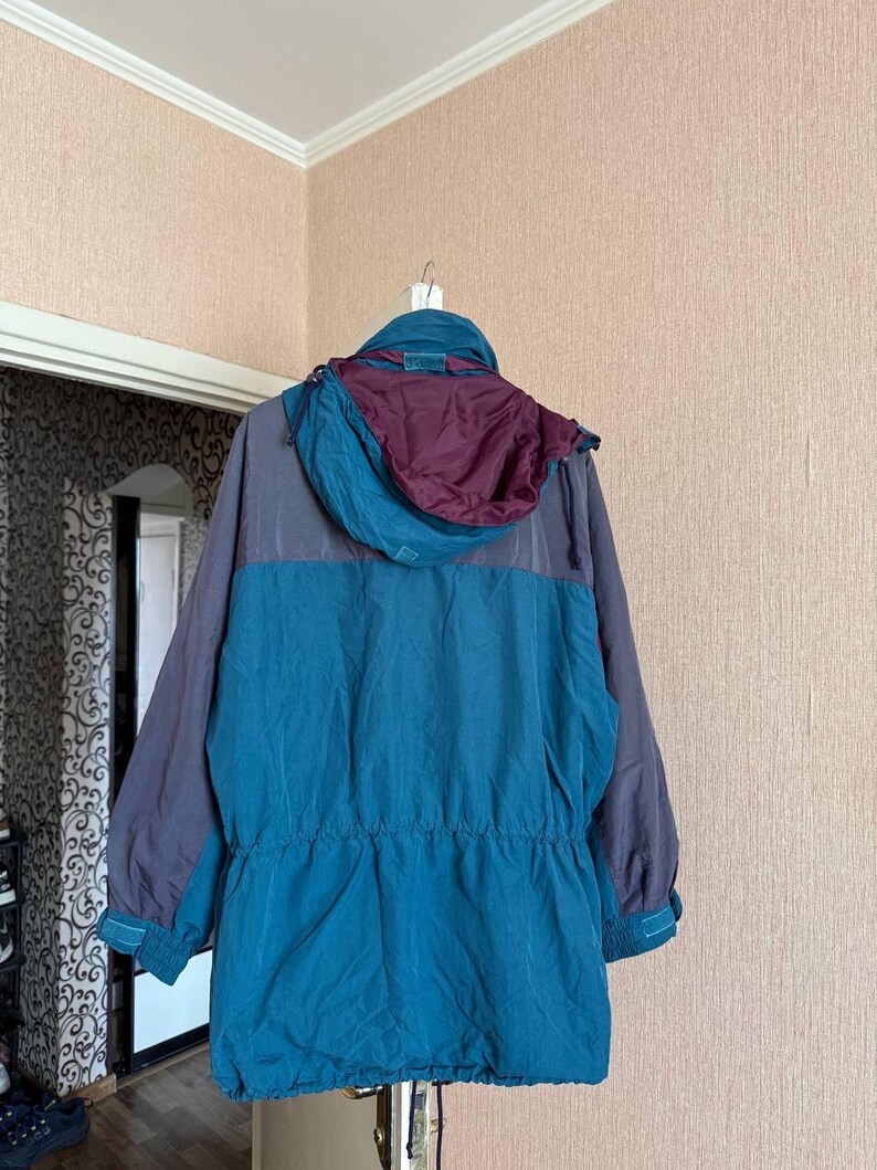 K&ouml;nnte beinhalten: Eine lang&auml;rmelige Jacke mit einem Color-Block-Design in Petrol und Dunkelgrau. Das Kleidungsst&uuml;ck verf&uuml;gt &uuml;ber eine Kapuze mit tiefburgunderrotem Innenfutter, eine Taille mit Kordelzug und verstellbare &Auml;rmelb&uuml;ndchen mit Klettverschl&uuml;ssen.