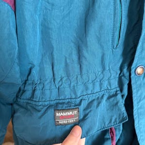 K&ouml;nnte beinhalten: Eine petrolfarbene Jacke mit violetten Akzenten, ausgestattet mit einer Brusttasche mit Rei&szlig;verschluss und einer Fronttasche mit Klappe. Ein schwarzer rechteckiger Aufn&auml;her auf der Klappentasche zeigt den Text MAMMUT SPORT und GORE-TEX.