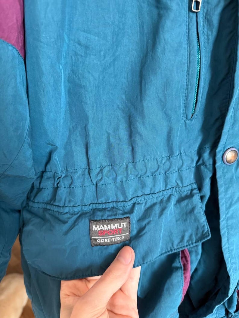 K&ouml;nnte beinhalten: Eine petrolfarbene Jacke mit violetten Akzenten, ausgestattet mit einer Brusttasche mit Rei&szlig;verschluss und einer Fronttasche mit Klappe. Ein schwarzer rechteckiger Aufn&auml;her auf der Klappentasche zeigt den Text MAMMUT SPORT und GORE-TEX.