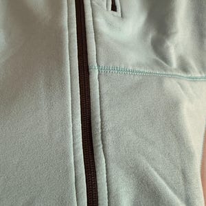 Puede incluir: Primer plano de una chaqueta azul claro con cremallera. La chaqueta tiene una cremallera marr&oacute;n oscuro y un peque&ntilde;o bolsillo. La tela tiene una textura suave y aterciopelada. Las costuras son de un tono azul m&aacute;s claro.