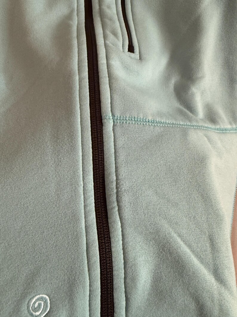 Puede incluir: Primer plano de una chaqueta azul claro con cremallera. La chaqueta tiene una cremallera marr&oacute;n oscuro y un peque&ntilde;o bolsillo. La tela tiene una textura suave y aterciopelada. Las costuras son de un tono azul m&aacute;s claro.