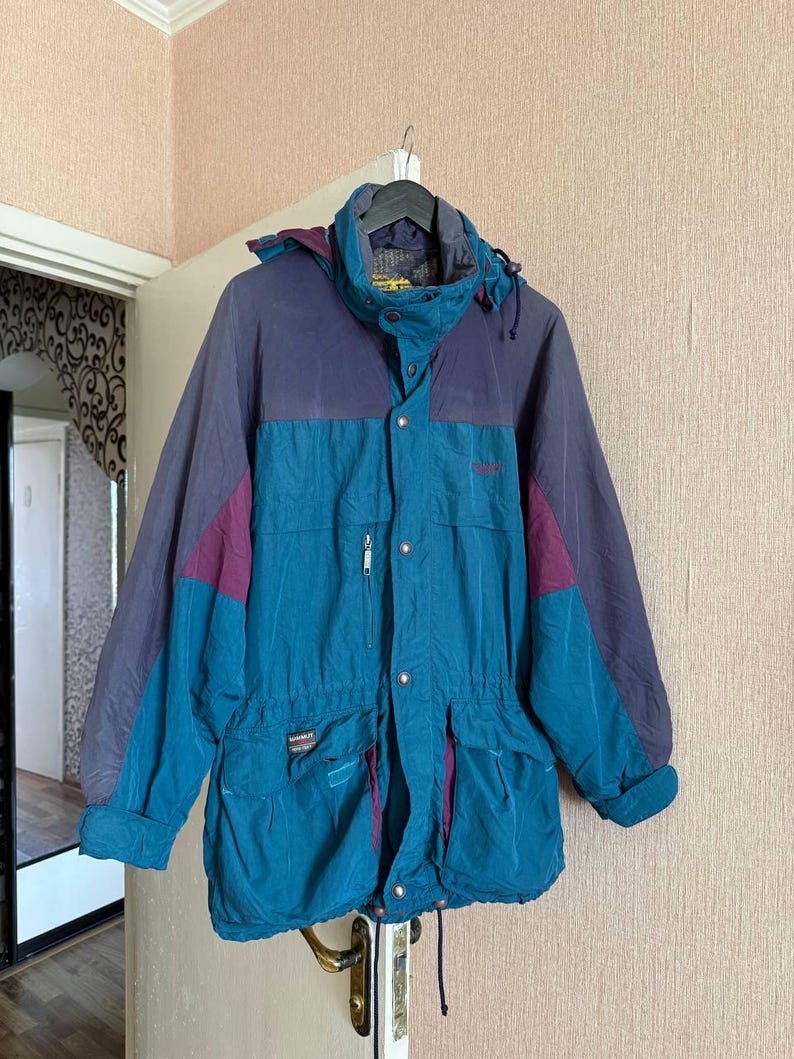 K&ouml;nnte beinhalten: Eine farblich abgesetzte Jacke mit einem t&uuml;rkisfarbenen Hauptteil und dunkelvioletten Eins&auml;tzen an Schultern und &Auml;rmeln. Die Jacke verf&uuml;gt &uuml;ber einen hohen Kragen, eine abnehmbare Kapuze, einen Frontrei&szlig;verschluss mit Druckkn&ouml;pfen und Taschen.