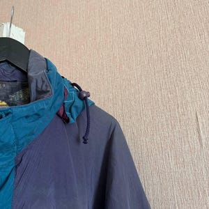 K&ouml;nnte beinhalten: Eine petrolfarbene und dunkelviolette Kapuzenjacke mit hohem Kragen, Kordelzug mit Kunststoffstopper und dunklem Innenfutter. Das Kleidungsst&uuml;ck ist mit farblich abgesetzten Paneelen in kontrastierenden Blau- und Violettt&ouml;nen gestaltet.