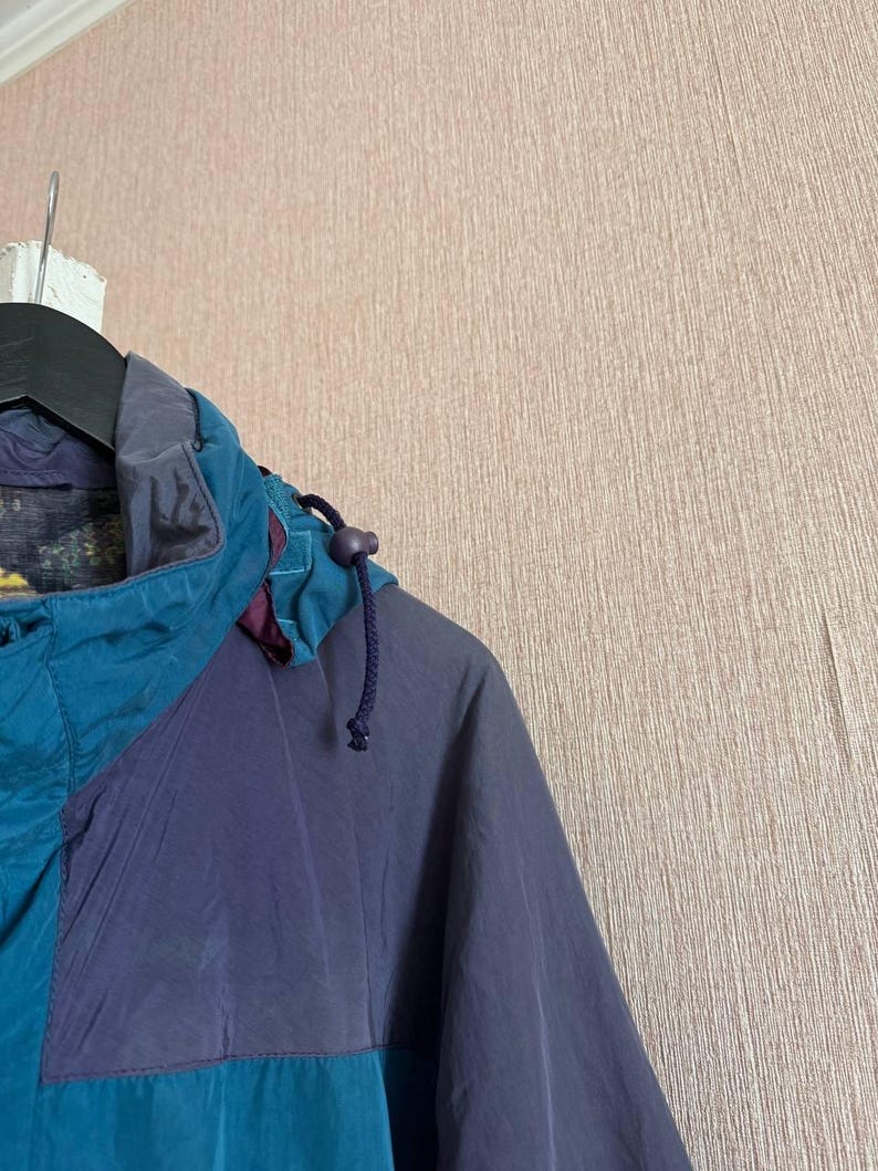 K&ouml;nnte beinhalten: Eine petrolfarbene und dunkelviolette Kapuzenjacke mit hohem Kragen, Kordelzug mit Kunststoffstopper und dunklem Innenfutter. Das Kleidungsst&uuml;ck ist mit farblich abgesetzten Paneelen in kontrastierenden Blau- und Violettt&ouml;nen gestaltet.