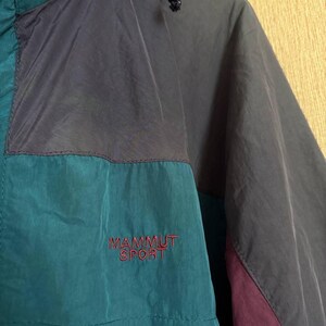 K&ouml;nnte beinhalten: Eine farblich abgesetzte Windjacke mit Eins&auml;tzen in Petrol, Dunkelgrau und Dunkelviolett. Die Jacke verf&uuml;gt &uuml;ber eine Kapuze mit Kordelzug und den in Rot auf die Brust gestickten Schriftzug MAMMUT SPORT. Der Stoff hat eine matte, leicht strukturierte Oberfl&auml;che.