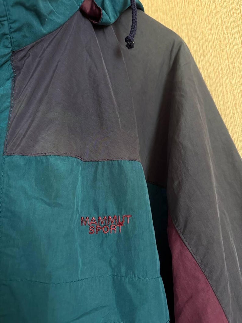 K&ouml;nnte beinhalten: Eine farblich abgesetzte Windjacke mit Eins&auml;tzen in Petrol, Dunkelgrau und Dunkelviolett. Die Jacke verf&uuml;gt &uuml;ber eine Kapuze mit Kordelzug und den in Rot auf die Brust gestickten Schriftzug MAMMUT SPORT. Der Stoff hat eine matte, leicht strukturierte Oberfl&auml;che.