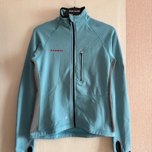 Puede incluir: Chaqueta azul claro con cremallera completa, cuello alto y mangas largas. La chaqueta tiene una cremallera negra, un peque&ntilde;o bolsillo en el pecho y paneles laterales grises. El nombre de la marca "MAMMUT" est&aacute; impreso en rojo.