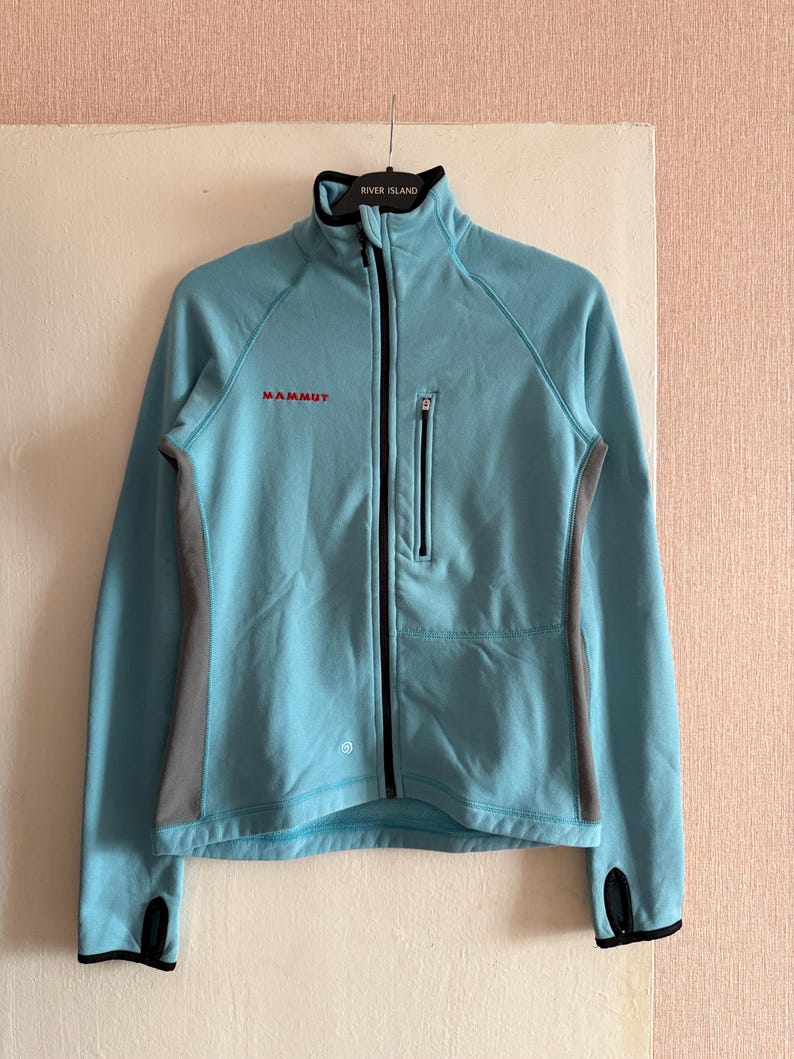Puede incluir: Chaqueta azul claro con cremallera completa, cuello alto y mangas largas. La chaqueta tiene una cremallera negra, un peque&ntilde;o bolsillo en el pecho y paneles laterales grises. El nombre de la marca "MAMMUT" est&aacute; impreso en rojo.