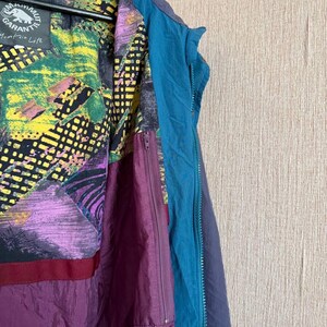 K&ouml;nnte beinhalten: Eine Jacke mit dunkelblauer Au&szlig;enseite und t&uuml;rkisfarbener Rei&szlig;verschlussleiste. Das Innenfutter zeigt ein lebhaftes, mehrfarbiges abstraktes Muster in Gelb, Gr&uuml;n, Lila und Schwarz. Ein Etikett tr&auml;gt die Aufschrift Mammut Garantie Mountain Life.
