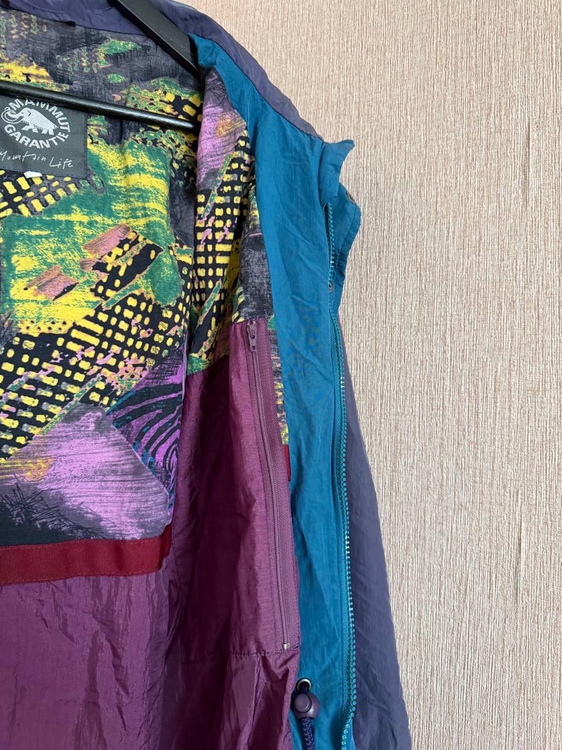 K&ouml;nnte beinhalten: Eine Jacke mit dunkelblauer Au&szlig;enseite und t&uuml;rkisfarbener Rei&szlig;verschlussleiste. Das Innenfutter zeigt ein lebhaftes, mehrfarbiges abstraktes Muster in Gelb, Gr&uuml;n, Lila und Schwarz. Ein Etikett tr&auml;gt die Aufschrift Mammut Garantie Mountain Life.
