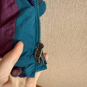 K&ouml;nnte beinhalten: Eine petrolfarbene und violette Jacke mit einem petrolfarbenen Rei&szlig;verschluss und zwei silbernen YKK Vislon-Rei&szlig;verschlussschiebern. Die Jacke besteht aus einem glatten, leichten Material und hat eine dunkelviolette Kordel am Kragenbereich.