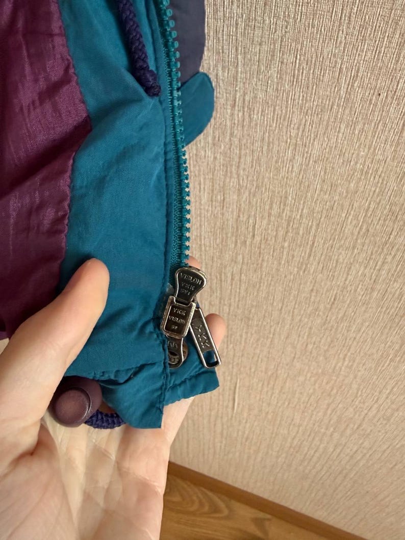 K&ouml;nnte beinhalten: Eine petrolfarbene und violette Jacke mit einem petrolfarbenen Rei&szlig;verschluss und zwei silbernen YKK Vislon-Rei&szlig;verschlussschiebern. Die Jacke besteht aus einem glatten, leichten Material und hat eine dunkelviolette Kordel am Kragenbereich.