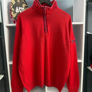 May include: A bright red quarter-zip pullover sweater featuring a high collar with a dark blue patterned trim along the zipper placket. The sweater includes a small zippered pocket on the left sleeve and ribbed cuffs and hem for a structured fit.