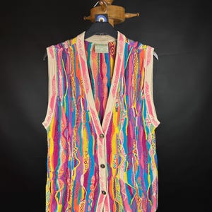 Puede incluir: Un chaleco multicolor con botones y cuello en V. El chaleco presenta una vibrante mezcla de rayas verticales rosas, azules, amarillas y naranjas. Los bordes y el cuello son de color beige neutro. El chaleco se exhibe en una percha de madera.