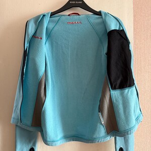 Puede incluir: Chaqueta azul claro con detalles en negro y gris, mostrada abierta. La chaqueta presenta un panel de malla negra en el lado derecho, un panel gris en el izquierdo y la marca "Prada" en rojo. Tiene orificios para los pulgares y una cremallera.