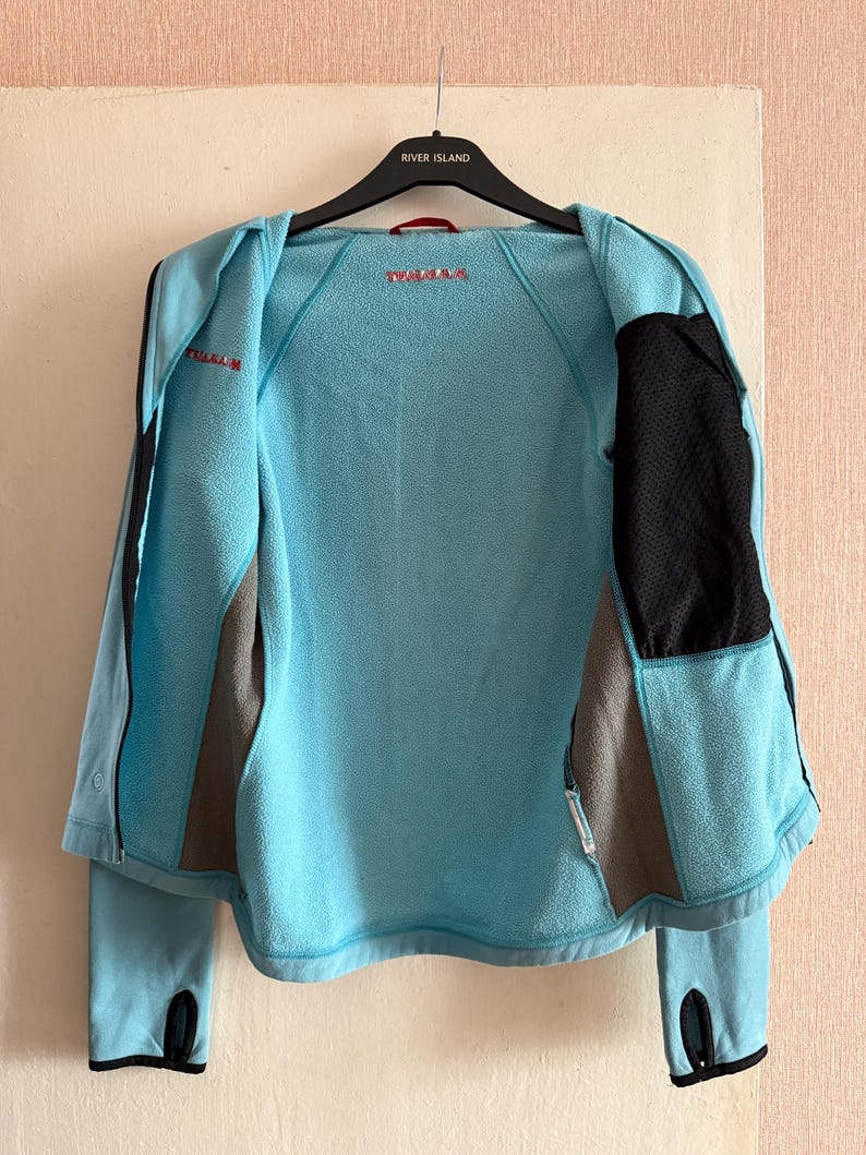 Puede incluir: Chaqueta azul claro con detalles en negro y gris, mostrada abierta. La chaqueta presenta un panel de malla negra en el lado derecho, un panel gris en el izquierdo y la marca "Prada" en rojo. Tiene orificios para los pulgares y una cremallera.