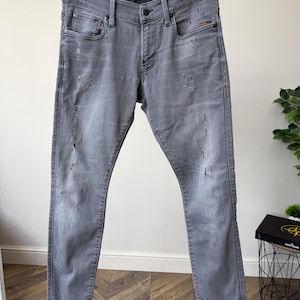 Könnte beinhalten: Graue Jeans aus Denim mit schmaler Passform, Knopfleiste und Used-Optik mit kleinen Rissen und Abriebstellen auf den Vorderteilen. Am Bund befindet sich ein kleines Etikett mit dem Text REVEND und der Stoff weist eine verwaschene Optik auf.