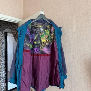 K&ouml;nnte beinhalten: Eine dunkelblaue und blaugr&uuml;ne Jacke mit einem mehrfarbigen Innenfutter, das abstrakte geometrische Muster in Gelb, Lila und Schwarz aufweist. Die Jacke hat ein einfarbiges dunkelblaues &Auml;u&szlig;eres, einen blaugr&uuml;nen Innenbesatz und einen einfarbigen violetten unteren Futterbereich.