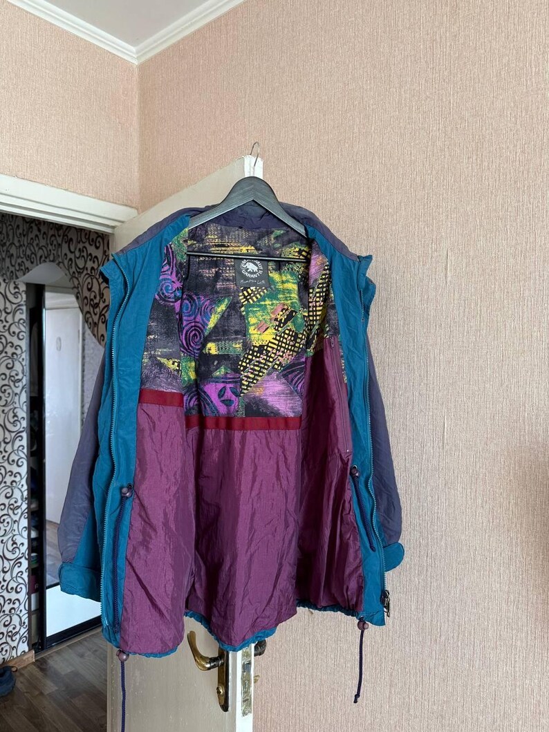 K&ouml;nnte beinhalten: Eine dunkelblaue und blaugr&uuml;ne Jacke mit einem mehrfarbigen Innenfutter, das abstrakte geometrische Muster in Gelb, Lila und Schwarz aufweist. Die Jacke hat ein einfarbiges dunkelblaues &Auml;u&szlig;eres, einen blaugr&uuml;nen Innenbesatz und einen einfarbigen violetten unteren Futterbereich.