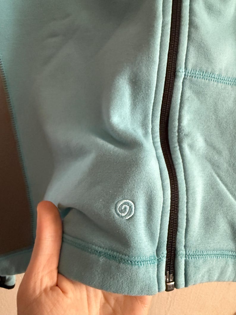 Puede incluir: Primer plano de una chaqueta azul claro con cremallera. La chaqueta tiene una cremallera negra y un peque&ntilde;o logotipo de espiral bordado en el pecho. La tela parece suave y texturizada.