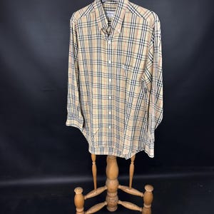 May include: A long-sleeved button-down shirt with a beige, black, red, and white plaid pattern. The shirt is displayed on a wooden hanger and stand against a black backdrop. The shirt has a small chest pocket. The shirt is made of a woven fabric.