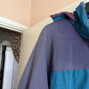 K&ouml;nnte beinhalten: Eine farblich abgesetzte Windjacke mit einem violetten Oberteil und einem blaugr&uuml;nen Unterteil. Die Jacke verf&uuml;gt &uuml;ber eine Kapuze im violett-blaugr&uuml;nen Design, lange &Auml;rmel und eine glatte, leichte Stoffkonstruktion mit einer sichtbaren Naht.