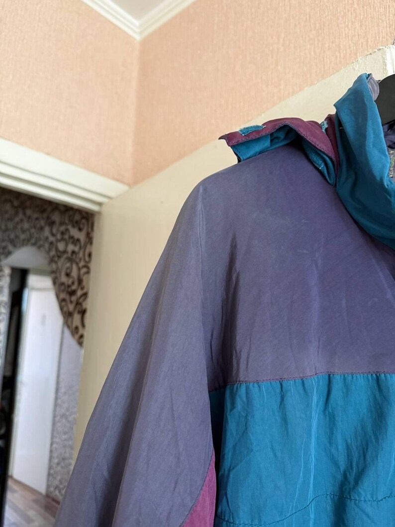 K&ouml;nnte beinhalten: Eine farblich abgesetzte Windjacke mit einem violetten Oberteil und einem blaugr&uuml;nen Unterteil. Die Jacke verf&uuml;gt &uuml;ber eine Kapuze im violett-blaugr&uuml;nen Design, lange &Auml;rmel und eine glatte, leichte Stoffkonstruktion mit einer sichtbaren Naht.