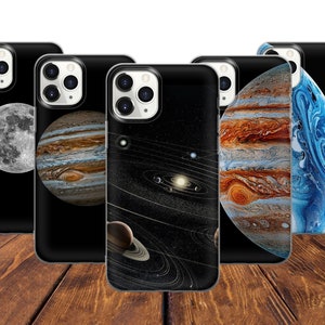 May include: Five different phone cases with a space theme. Each case features a different planet, including a moon, Jupiter, Saturn, and a blue nebula. The cases are all black with a white outline around the planets.