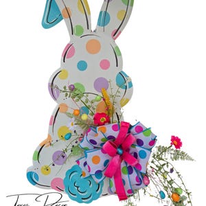 May include: A white wooden bunny cutout with colorful polka dots, decorated with a pink and white polka dot bow, green and yellow floral sprigs, and colorful Easter eggs. The bunny is standing on a white surface. Texas Decor By Nana is written in black at the bottom of the image.