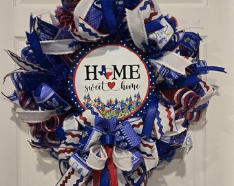 Peut inclure: Une couronne rouge, blanche et bleue avec une pancarte qui dit "Home sweet home" avec une silhouette du Texas et un cœur. La couronne est décorée de rubans et de filet bleu, rouge et blanc.