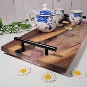 Peut inclure: Un plateau de service en bois avec une poignée noire et un incrustation de résine rose avec des fleurs pressées. Le plateau est décoré d'un motif ondulé et a une finition en bois naturel.