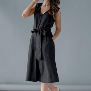 Op de afbeelding: Een mouwloze zwarte jumpsuit met een V-hals en knoopsluiting. De jumpsuit heeft een bijpassende stoffen riem die in de taille is vastgemaakt en een culotte met wijde pijpen. Zakken zijn zichtbaar aan de zijkanten.