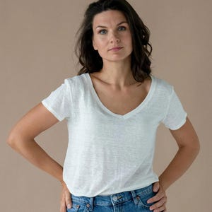 以下が含まれることがあります： 白いVネックTシャツとブルージーンズ。半袖シャツは軽量の生地で作られています。モデルはニュートラルな背景を背景に、腰に手を当てて立っています。