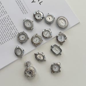 Puede incluir: Una colección de pequeñas esferas de reloj plateadas en varias formas, incluyendo redondas, ovaladas y en forma de corazón. Algunas tienen biseles decorativos y números romanos. Los relojes están dispuestos sobre una superficie blanca.