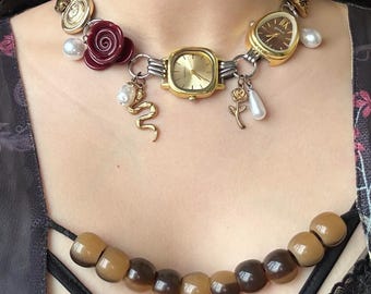 Pode incluir: Um colar único com mostradores de relógios reaproveitados, metal dourado e pingentes decorativos. O colar inclui um mostrador de relógio quadrado dourado, uma rosa bordô, pérolas falsas e um pingente de cobra dourada. A parte inferior do colar é feita de contas marrons.