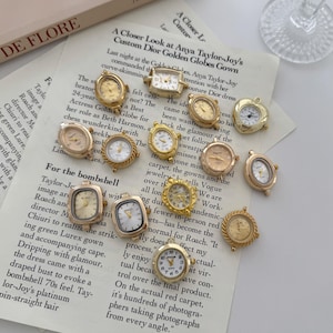 May include: A collection of small, gold-toned watch charms in various shapes, including round, oval, and square. The watch faces feature Roman numerals and delicate hands, resting on a printed paper background with text.