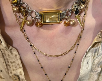 May include: A multi-strand necklace featuring a gold watch face, various charms, and multiple chains. The necklace includes gold and silver-toned metal elements, with accents of black beads and a snake pendant. The watch face is rectangular with a gold frame.