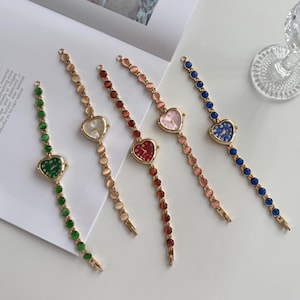 Può includere: Cinque orologi a forma di cuore con cinturini in metallo dorato e dettagli in pietre colorate. I quadranti degli orologi sono verdi, bianchi, rossi, rosa e blu. I cinturini presentano pietre colorate abbinate. Gli orologi sono esposti su una superficie bianca.